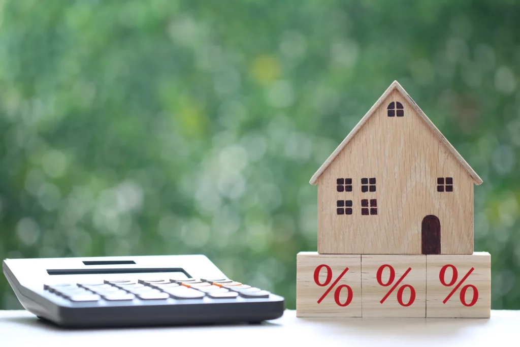 Une maison miniature en bois posée sur des blocs marqués de pourcentages, avec une calculatrice à proximité, illustrant le concept des taux d'intérêt ou des calculs financiers liés à l'immobilier.