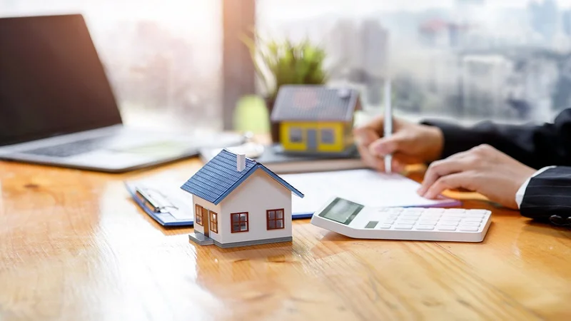 Scène représentant le calcul des frais liés à un prêt immobilier, avec une maison miniature, une calculatrice et des documents sur un bureau.