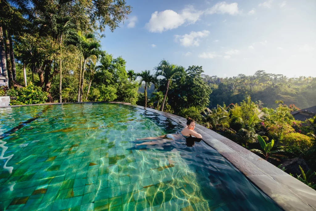 Une femme se détend dans une piscine à débordement offrant une vue panoramique sur une jungle luxuriante et un paysage tropical sous un ciel ensoleillé.