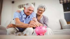 L'image montre un couple de retraités souriants ajoutant une pièce dans une tirelire rose, symbolisant l'épargne et la préparation financière pour une retraite sereine.