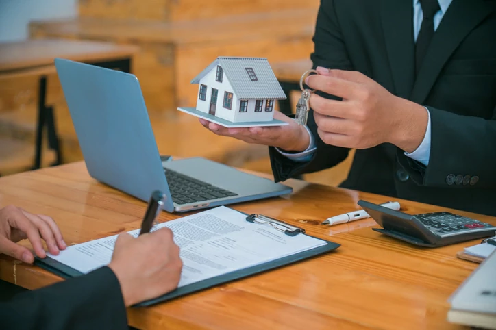 indivision ou SCI : personne tenant une petite maison et une clé devant une autre personne signant un document à coté d'un ordinateur portable