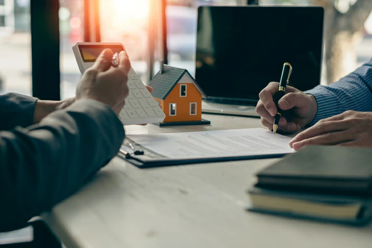 prêt différé : personne écrivant sur un bloc-note devant une autre personne tenant une calculatrice à coté d'une petite maison