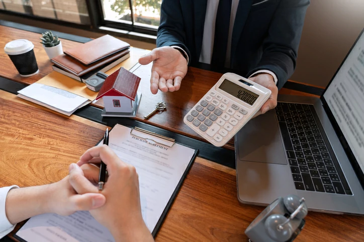 comptabilité des sci : personne tenant une calculatrice devant une personne observant un document à coté d'une petite maison