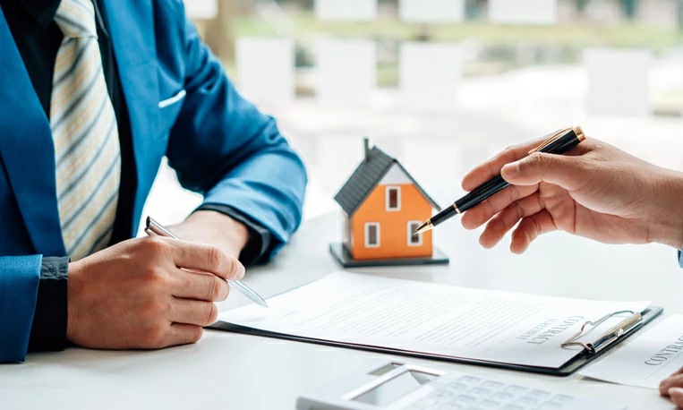 sci nu propriété : personne pointant un document en papier avec un stylo devant une autre personne qui se prépare à signer le document à coté d'une petite maison.