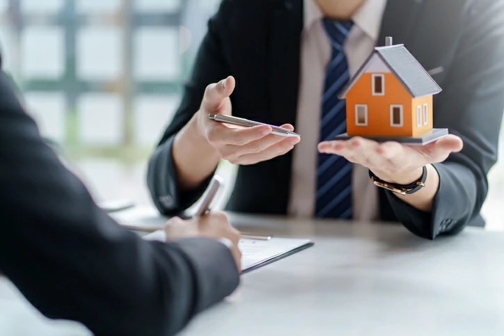 sci nu propriété : personne tenant une petite maison dans la main gauche et un stylo dans la main droite devant une personne qui signe un document en papier